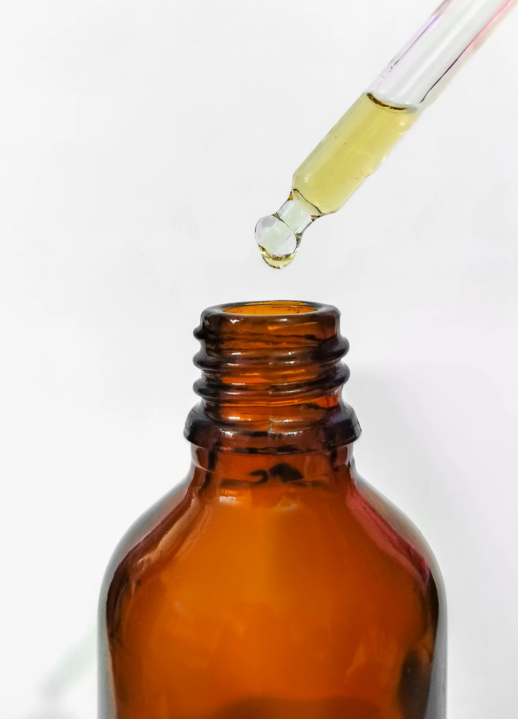 A glass pipette releasing a single drop of golden oil into the neck of an amber bottle against a plain white ground