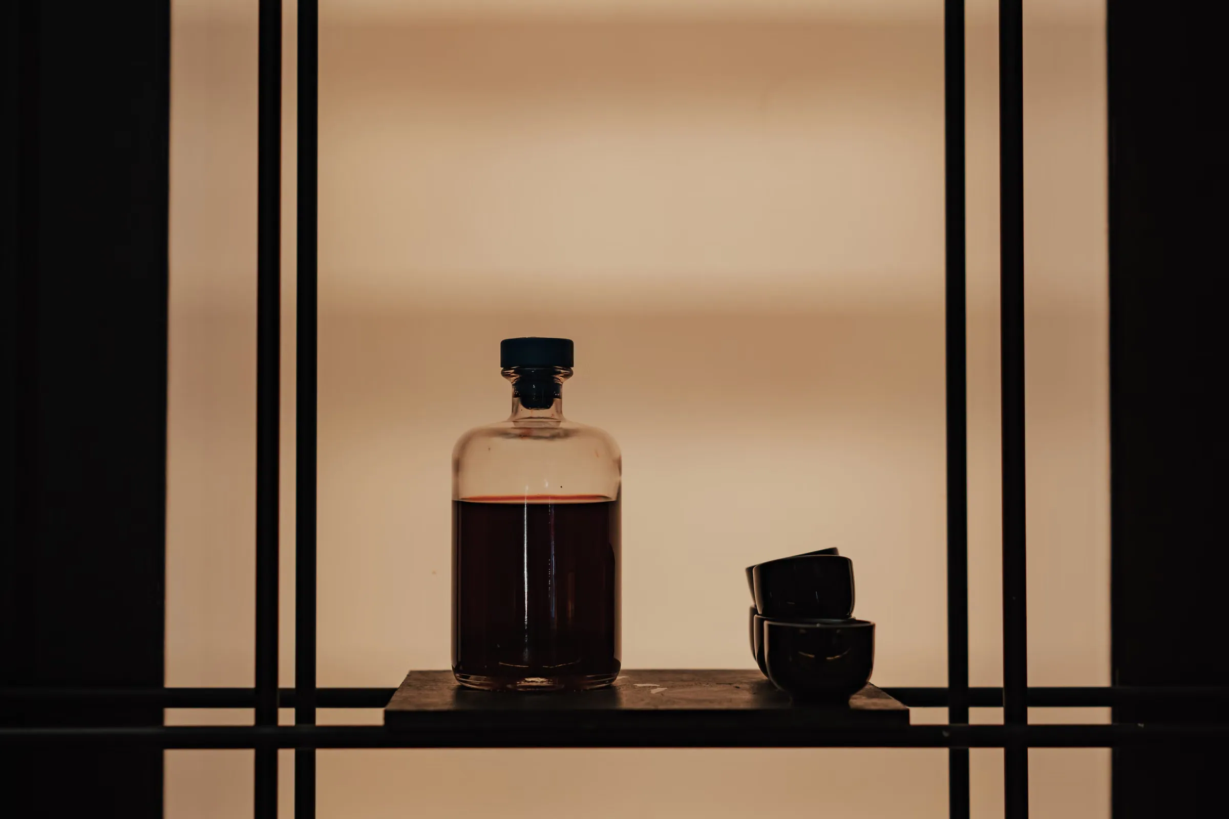 A dark glass bottle and two small ceramic cups on a black shelf against a plain warm wall, in low even light