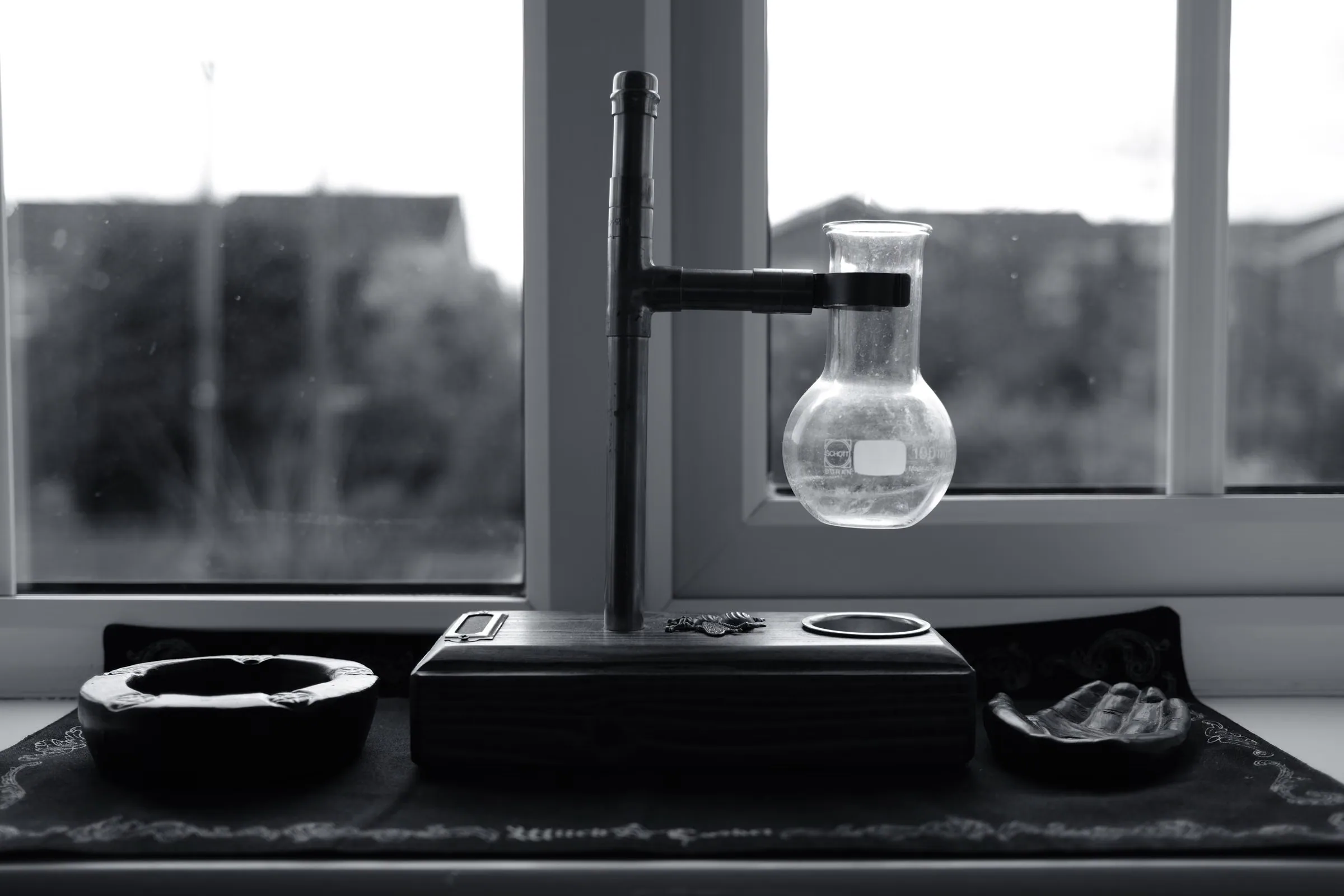 Black and white still life of a round glass flask standing on a balance on a windowsill, city rooftops beyond