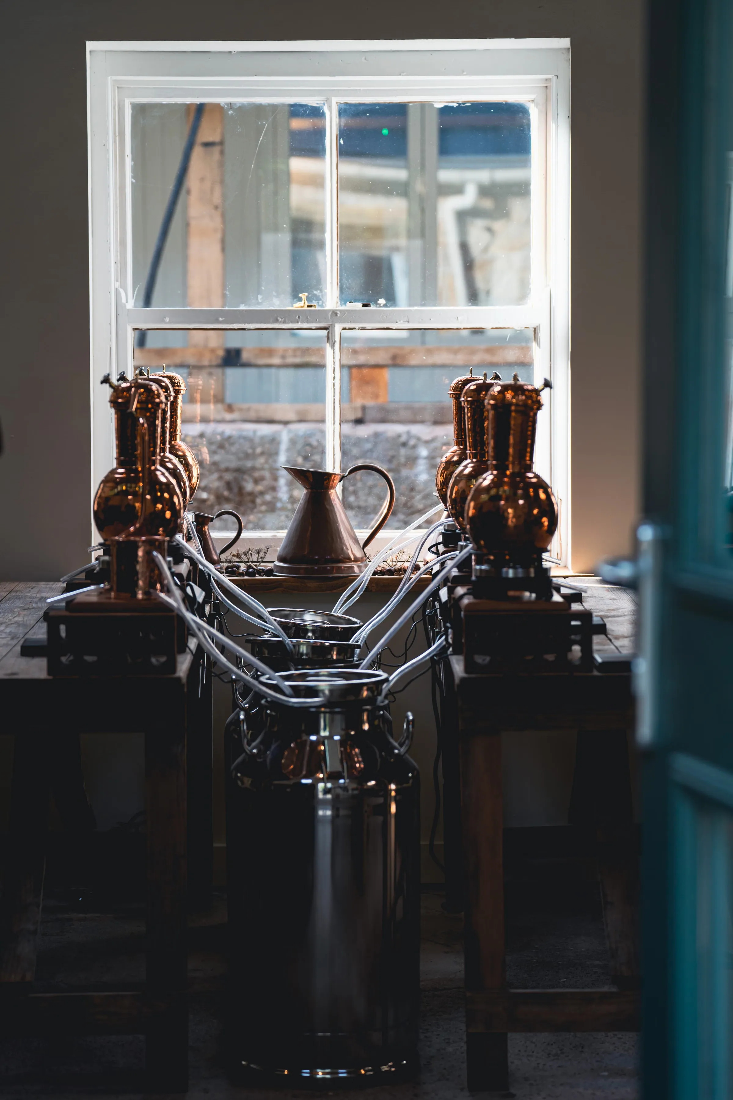 Small copper distillation heads on a bench in front of a bright window, silhouetted against daylight