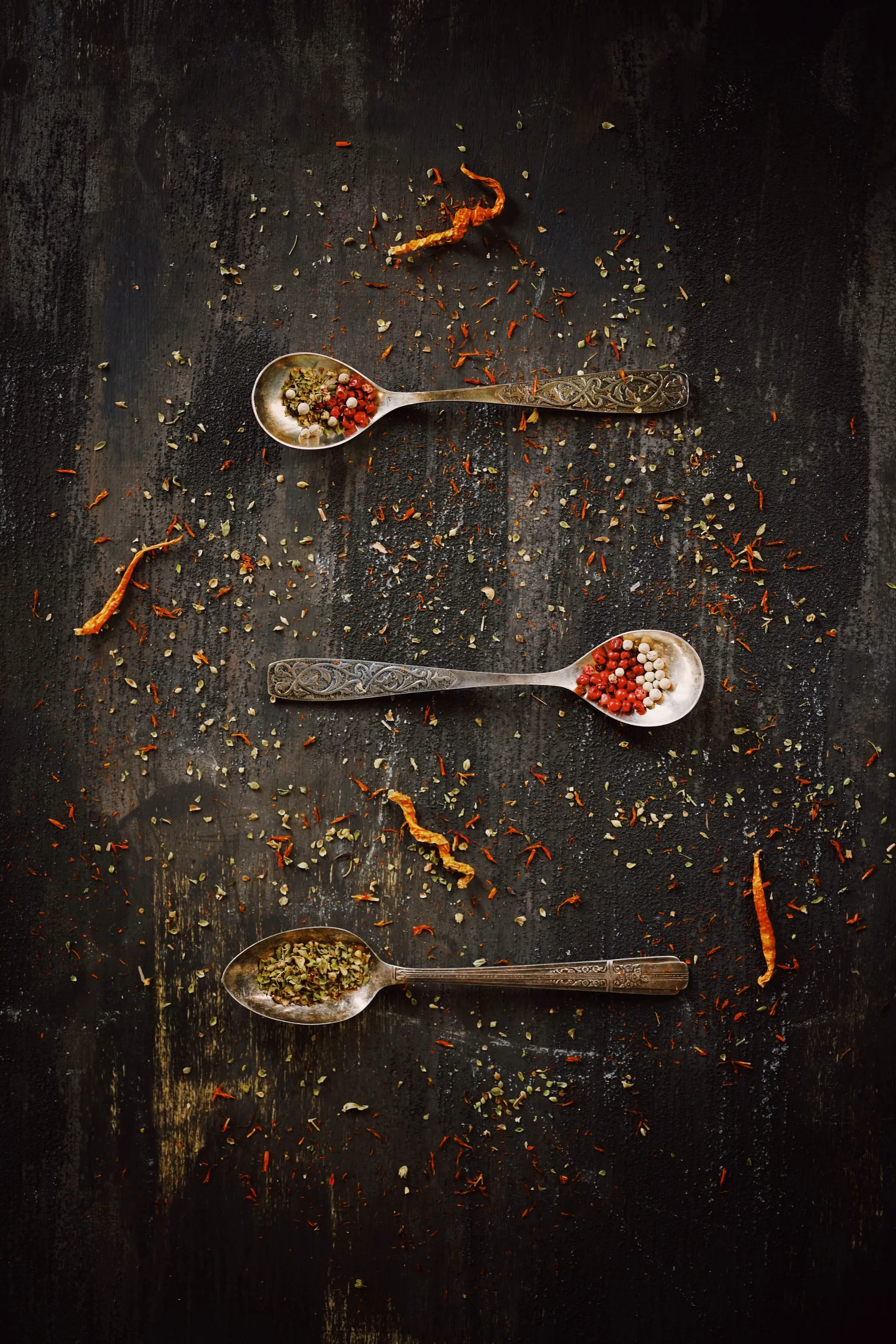 Engraved metal spoons holding resins and ground spice, scattered across near-black scorched wood
