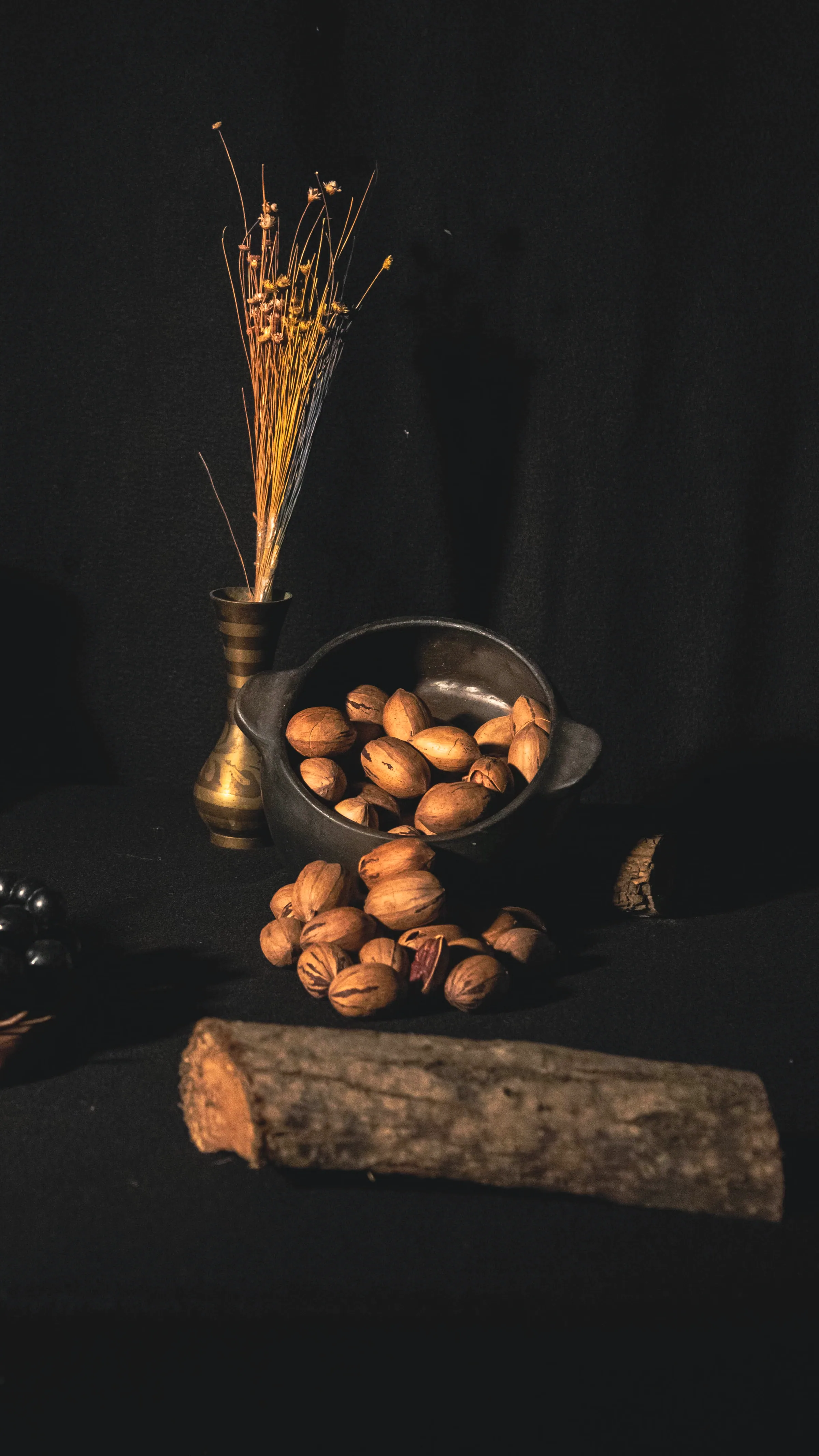 Pecans spilling from an iron bowl beside a brass vase of dried stems and a raw log, on black cloth
