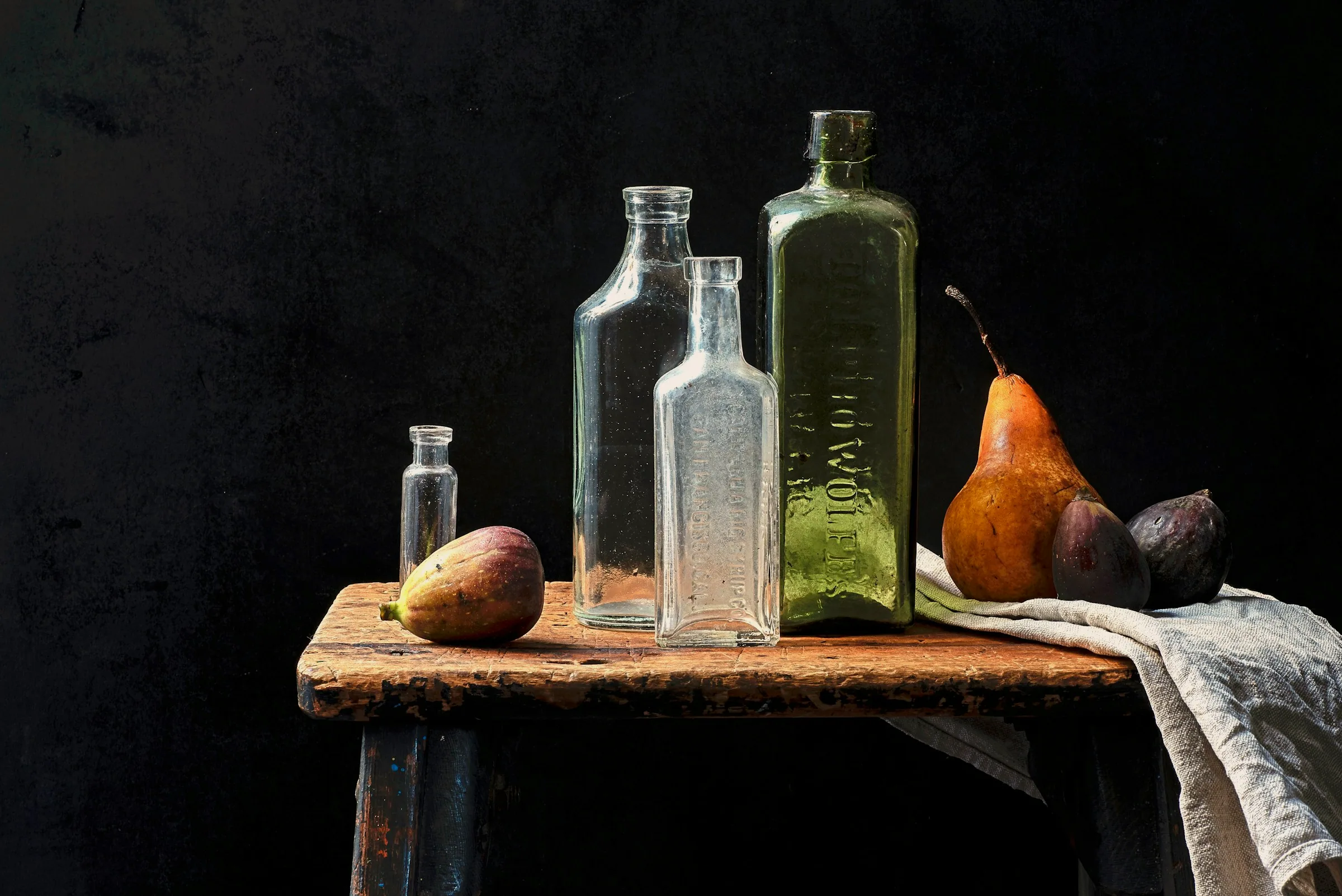 Antique glass bottles and pears on a worn wooden bench, lit from one side against a near-black wall