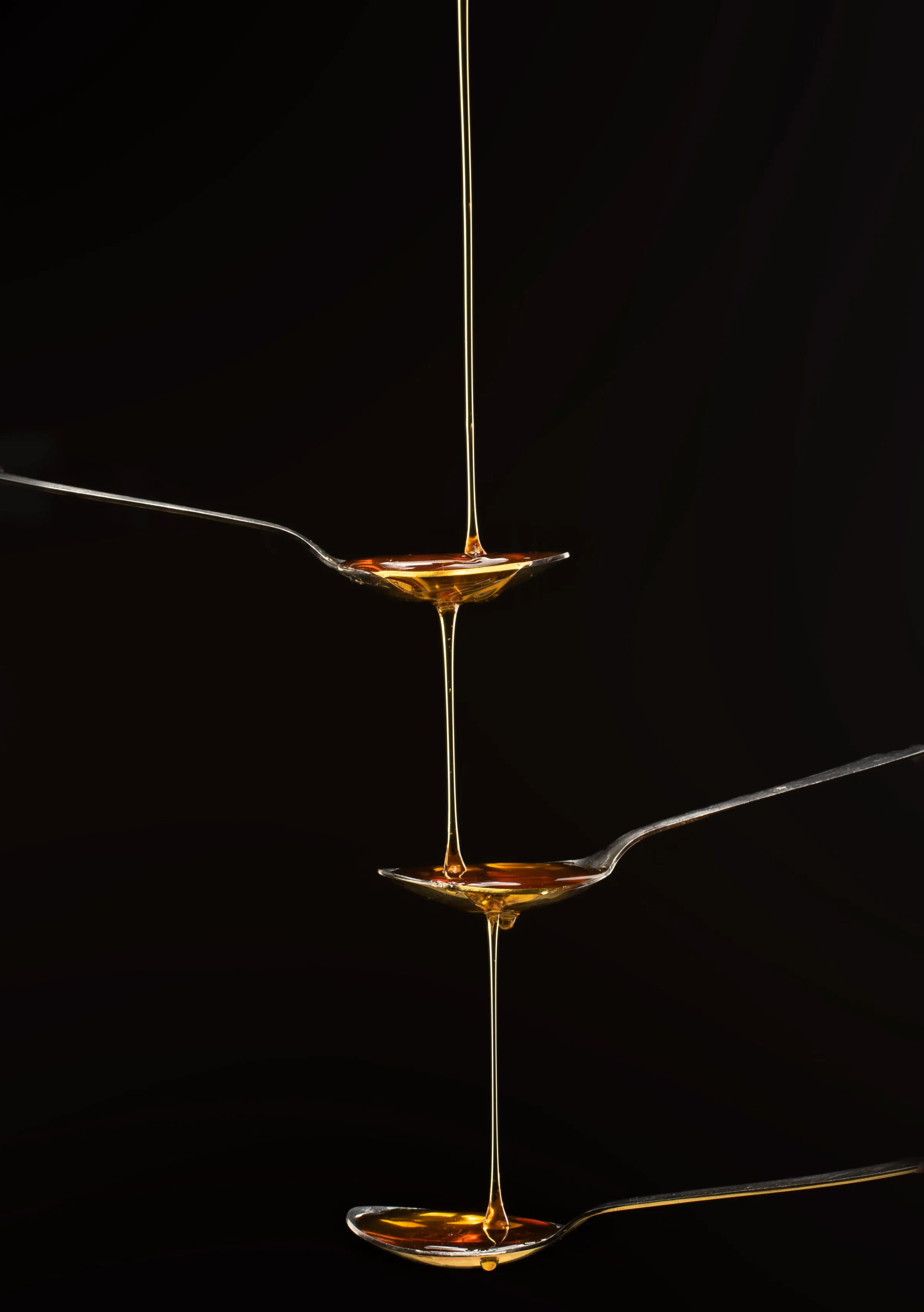 A golden thread of liquid falling through three suspended spoons against jet black