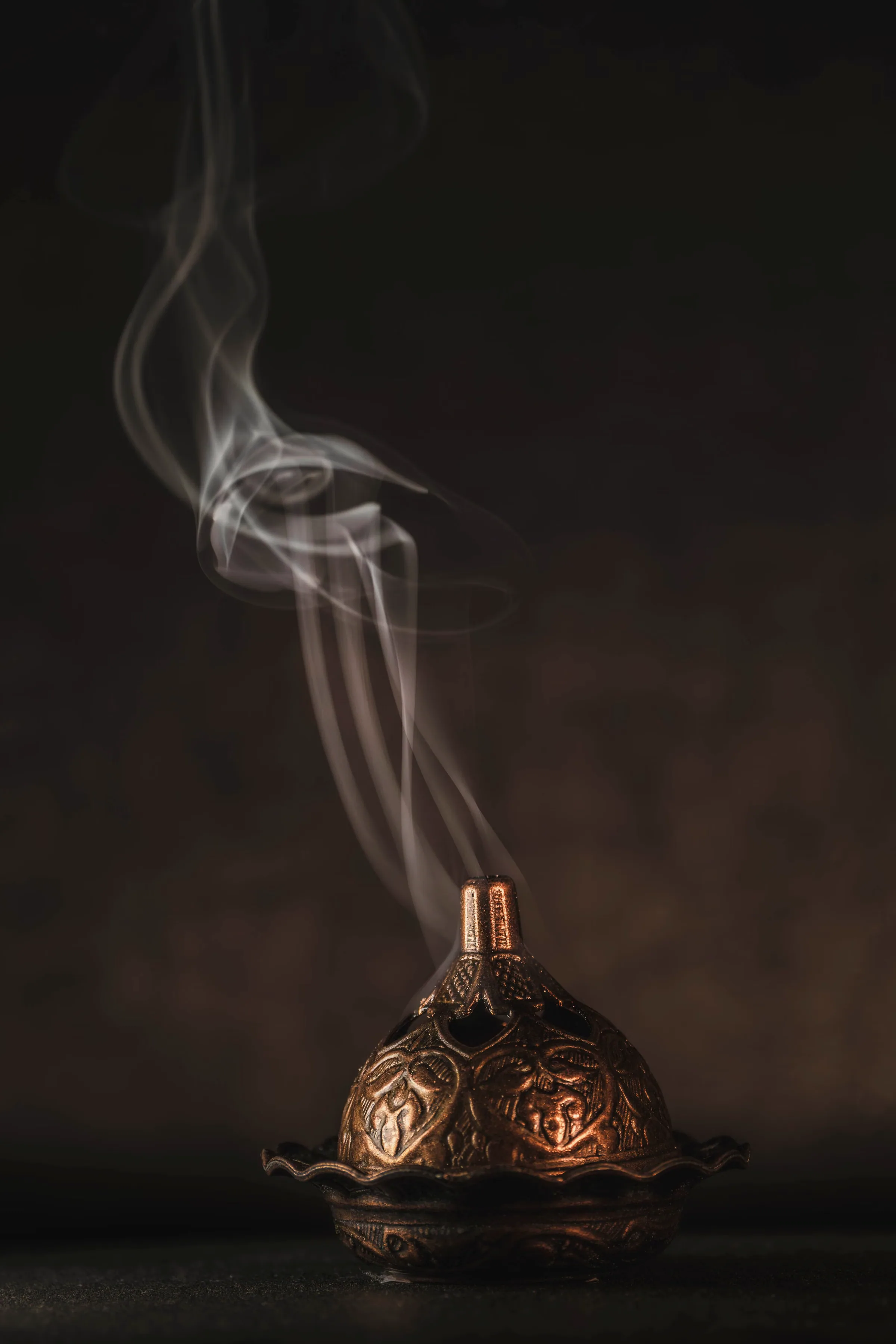 An engraved copper incense burner releasing one slow ribbon of smoke against an umber-black ground