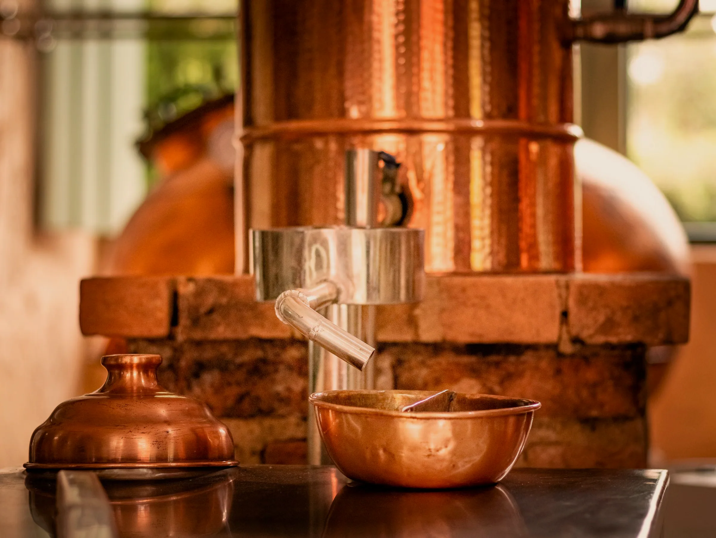 A hammered copper still on a brick hearth, its spout over a polished copper bowl, everything glowing amber