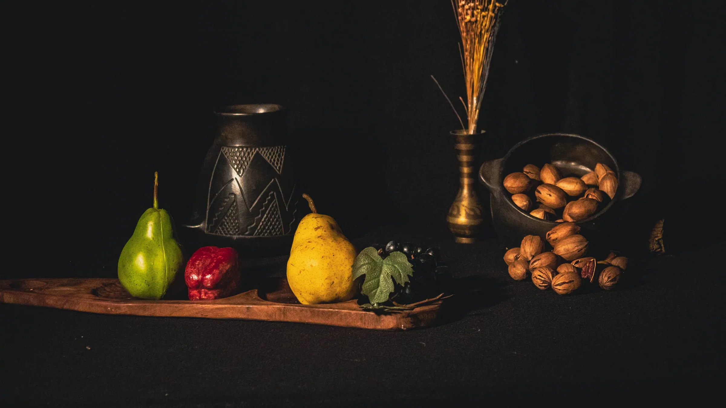 A still-life tableau of pears, figs, pecans, a brass vase and black vessels on a dark cloth