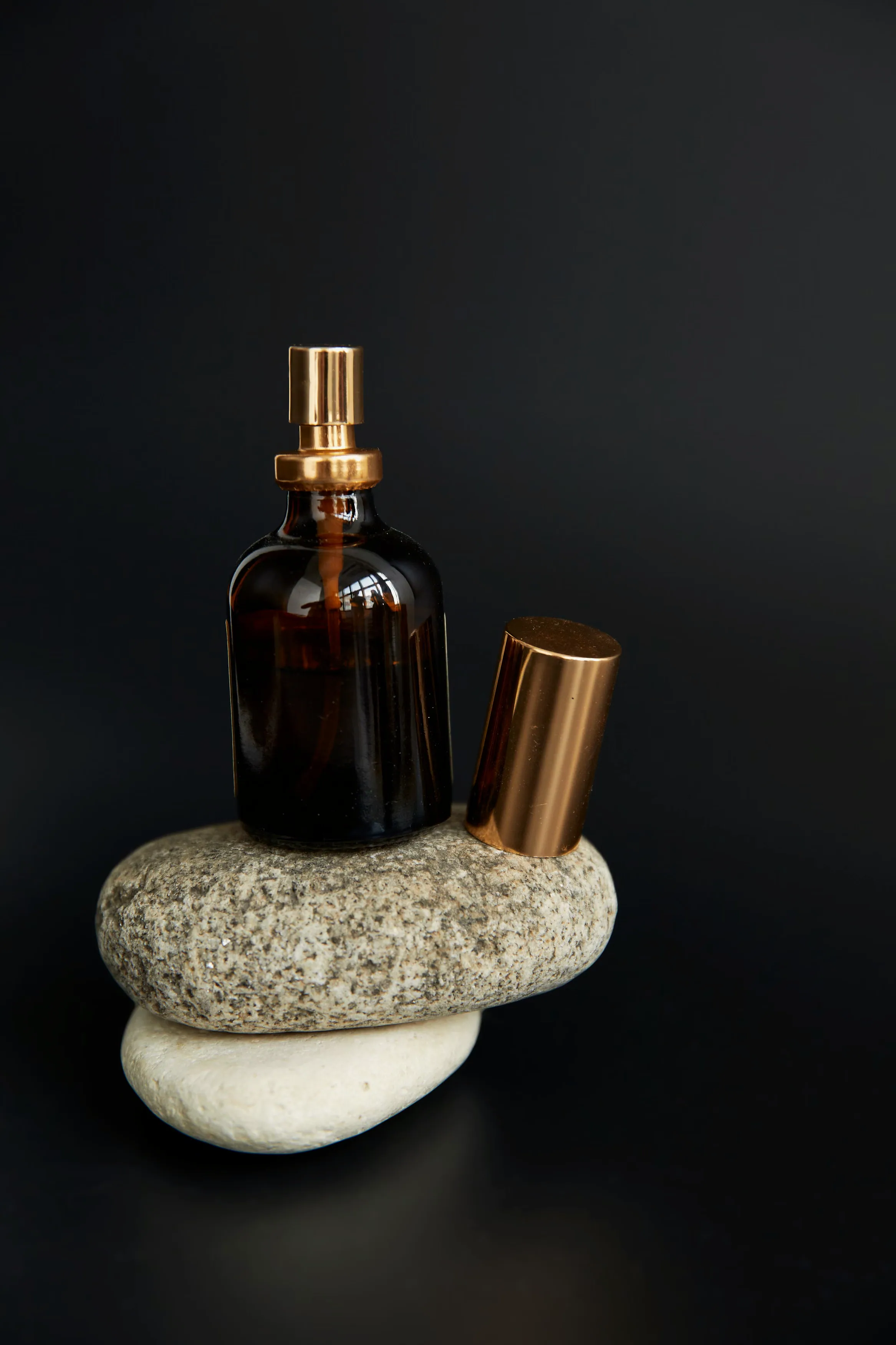 A single amber glass bottle with a copper atomizer cap standing on stacked stones against a black backdrop
